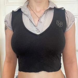 Svart croppad linnetopp med emblem - Svart croppad/avklippt linnetopp med v-ringning och ett litet broderat emblem på vänster sida. Toppen är ärmlös och har en enkel, stilren design som passar perfekt till lager-på-lager-stil. Mjuk och stretchig känsla. 