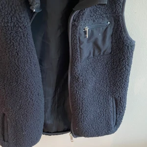Svart fleeceväst från 157, stl 140 - Mysig svart fleeceväst från 157 med dragkedja framtill och praktiska fickor, inklusive en bröstficka med dragkedja. Västen har en enkel och stilren design med mjukt teddyfoder som håller dig varm under kyliga dagar.