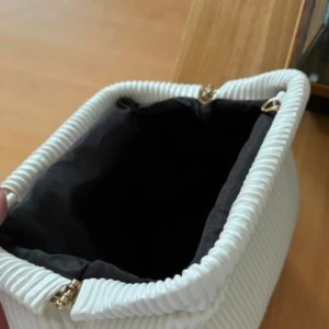 Vit clutch - Säljer en stilren vit kuvertväska med ribbad struktur och mjuk form. Väskan har guldfärgade detaljer och svart innerfoder. Perfekt för dig som gillar minimalistisk och modern design. Materialet är skinnimitation och väskan har en unik, veckad look.