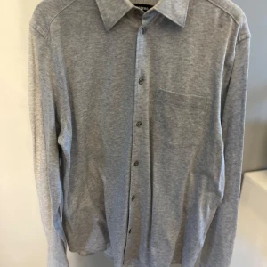 Ljusgrå skjorta med bröstficka - Säljer en stilren ljusgrå skjorta med klassisk krage och knäppning framtill. Skjortan har en bröstficka på vänster sida och långa ärmar. Perfekt för  dig som gillar en enkel och clean look.