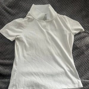 Vit ribbad polotopp - Säljer en vit ribbad polotopp med korta ärmar och klassisk krage. Toppen har en stilren och enkel design som passar perfekt till många olika outfits.