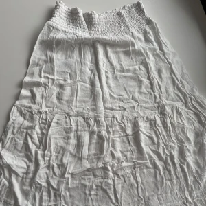 Vit långkjol med smock i midjan - Säljer en vit långkjol med smockad midja. Kjolen är luftig och har en lätt, flowy känsla som passar perfekt till varmare dagar. Enkel och stilren design som är lätt att matcha med olika toppar.