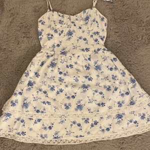 Vit blommig klänning från Hollister - Söt vit klänning men blå blommor från Hollister. Klänningen har spetsdetalj längst ner. Den skulle vara perfekt till skolavslutningen eller till midsommar, men den funkar perfekt att ha på vanliga sommar dagar.