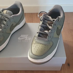 Nike Air Force 1 i olivgrönt - Säljer ett par Nike Air Force 1 i olivgrönt med grå detaljer och vit sula. Klassisk låg modell med snörning och rund tå. Perfekta sneakers för dig som gillar stilrena och tidlösa skor.