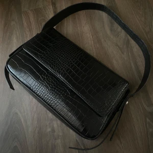 Svart axelväska i krokopräglat skinn - Säljer en svart axelväska i krokopräglat skinnimitation med bred axelrem. Väskan har ett stilrent och elegant utseende med lock och silverfärgad knappdetalj på sidan. Perfekt för dig som vill ha en trendig accessoar med lyxig känsla.
