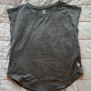 Mörkgrå träningst-shirt från SOC - Säljer en mörkgrå träningst-shirt från SOC med rund hals och diskret logga framtill. T-shirten är tillverkad i ett tekniskt material som andas och har en lätt, sportig känsla. Perfekt för aktiva dagar och träning.