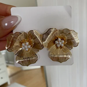Guldiga blomformade örhängen med pärlor oanvända  - Stora statement-örhängen i form av blommor i guldmetall med detaljerade blad och små vita pärlor i mitten. Perfekta för att lyfta vilken outfit som helst.