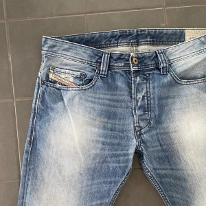 Blå raka jeans från Diesel - Säljer ett par klassiska blå jeans från Diesel med raka ben och snyggt slitna detaljer. Jeansen har fem fickor, synliga sömmar och en liten Diesel-logga på myntfickan. Perfekta för en avslappnad stil.