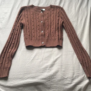 Brun croppad kofta från H&M  - Säljer en brun croppad kofta från H&M. Tröjan är stickad med kabelmönster, har långa ärmar och knappar framtill. Perfekt för lager-på-lager och enkel att styla till olika outfits. Säljer då den inte används. Ordpris. 179kr Nypris. 40kr