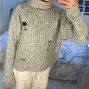 Grå stickad polotröja med slitningar - Säljer en oversized grå stickad polotröja med ribbstickning och flera slitna detaljer. Tröjan har lång ärm och hög polokrage, perfekt för en avslappnad och trendig look.