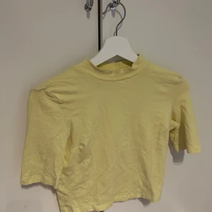 Ljusgul  t-shirt från Monki - Säljer en ljusgul croppad t-shirt från Monki. T-shirten har korta ärmar och en enkel, stilren design. Perfekt för varma dagar och enkel att matcha med jeans eller kjol. Aldrig använd. 