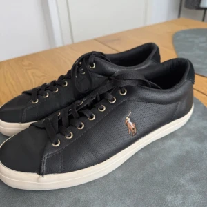 Polo Ralph Lauren sneakers – Svarta - Storlek: 41 Skick: SOM NYA – sparsamt använda Pris: 400 kr  Stilrena svarta sneakers från Polo Ralph Lauren med klassisk vit sula och broderad logotyp i brunt på sidan. Skorna är i mycket fint skick och endast försiktigt använda – det finns en liten skråma i hälen (syns inte vid användning).  🔹 Ovandel i läder 🔹 Bekvämt textilfoder 🔹 Slitstark gummisula 🔹 Broderad Polo-logotyp på sidan 🔹 Storlek 41 – normal passform