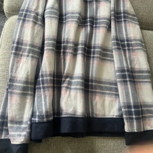 Rutig fleece tröja i blått och vitt - Mysig fleece tröja med rutigt mönster i blått, vitt och inslag av ljusrosa. Tröjan har långa ärmar och mörkblå muddar vid ärmslut och nederkant. Perfekt för kyliga dagar.