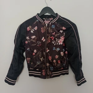 Svart bomberjacka med blommigt mönster från Urban Republic - Snygg svart bomberjacka från Urban Republic med färgglatt blommigt mönster och randiga muddar. Jackan har dragkedja framtill och långa ärmar. Perfekt för dig som vill sticka ut med en cool stil. (Barn jacka)