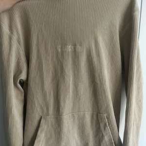 Beige ribbad långärmad tröja från Szerh - Säljer en beige långärmad tröja från Szerh med diskreta vertikala ränder och broderad logga på bröstet. Tröjan har en ficka framtill och är tillverkad i mjukt material som passar perfekt till vardags.