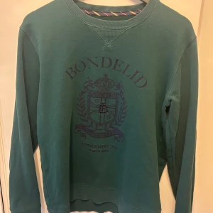 Grön sweatshirt från Bondelid - Säljer en grön sweatshirt från Bondelid med tryckt logga och emblem på bröstet. Tröjan har rund halsringning och långa ärmar. Perfekt för en avslappnad och stilren look.