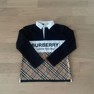 Burberry pikétröja med klassiskt rutmönster - Säljer en snygg pikétröja från Burberry med vit krage, svarta ärmar och överdel samt klassiskt beige rutmönster nertill. Stor logga framtill och knäppning vid halsen. Perfekt för dig som gillar stilrena märkeskläder.
