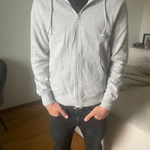 Grå zip-hoodie - Säljer en enkel grå hoodie med dragkedja och huva. Jackan har snörning vid huvan och två fickor framtill. Perfekt för en avslappnad stil och passar till det mesta.