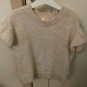 Beige stickad tröja med volangärmar från Gina Tricot - Superfin beige stickad tröja från Gina Tricot med korta volangärmar och bred ribbad kant vid hals, ärmslut och nederkant. Perfekt för lager på lager och ger en söt och trendig look.