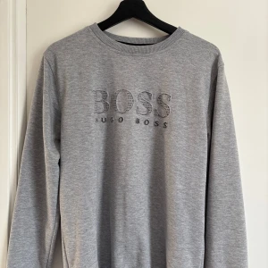 Grå sweatshirt från Hugo Boss - Säljer en stilren grå sweatshirt från Hugo Boss med broderad logga på bröstet. Tröjan har rund halsringning och långa ärmar. Perfekt för en avslappnad och snygg look.
