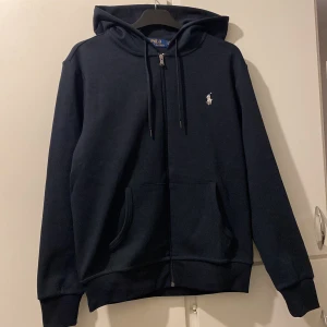 Ralph Lauren kofta - Snygg mörkblå hoodie från Polo Ralph Lauren med dragkedja och klassisk logga på bröstet. Jackan har huva med dragsko och två fickor framtill. Perfekt för en avslappnad stil.