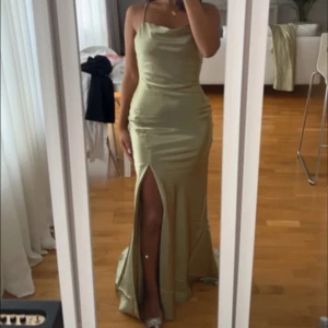 Champagnefärgad långklänning med slits - Säljer en elegant långklänning i champagnefärgat satinmaterial. Klänningen har smala axelband, figurnära passform och en hög slits framtill. Öppen rygg med tunna band som korsas. Perfekt för festliga tillfällen.