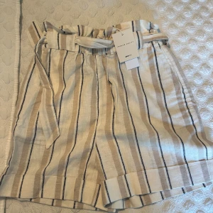 Randiga shorts med bälte från Gina Tricot - Snygga beige och vita shorts med bruna och svarta ränder från Gina Tricot. Modellen har hög midja, knytskärp och uppvikta benslut. Perfekta för varma dagar och ger en avslappnad men trendig look.