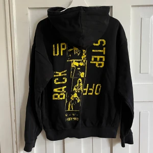 Svart hoodie med gult tryck från Stay - Säljer en svart hoodie från Stay, med tryck i gult på ryggen. Hoodien har känguruficka och huva med dragsko.