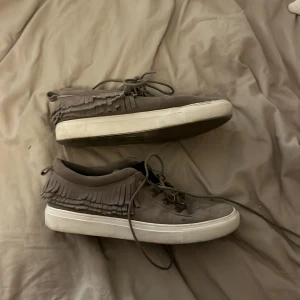 Grå mockasneakers med fransar från Kurt Geiger - Säljer ett par grå sneakers från Kurt Geiger i mocka med snygga fransdetaljer runt ankeln. Skorna har snörning och en vit platt sula som ger en stilren look. Perfekta för dig som vill ha något unikt och trendigt i garderoben