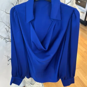 Blusen har lång ärm och är tillverkad i ett glansig - Säljer en blå blus  Blusen har lång ärm och är tillverkad i ett glansigt, mjukt material som ger en elegant känsla.
