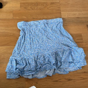 Blå blommig kjol från Lindex - Söt axelbandslös klänning från Lindex i ljusblått med vitt blommönster och volang nertill. Perfekt för varma dagar och har en luftig passform. Klänningen är i barnstorlek 146/152.