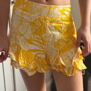 Gula och vita lågmidjade volangshorts - Säljer ett par lågmidjade gula och vita shorts med volangdetaljer längst ner. Perfekta för sommaren och har en lös passform. Materialet känns lätt och luftigt.
