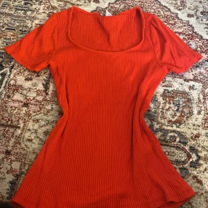 Ribbad röd topp med kort ärm - Säljer en ribbad röd topp med korta ärmar och rundad, djup urringning. Toppen har en figurnära passform och är perfekt för att lyfta en enkel outfit med lite färg.