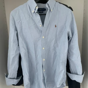 Ljusblå skjorta från Ralph Lauren - Klassisk ljusblå skjorta från Ralph Lauren med button-down krage och broderad logga på bröstet. Skjortan har långa ärmar och vita knappar framtill. Perfekt för en stilren och tidlös look. Kan tyvärr ej lösa bild me den på pga för liten