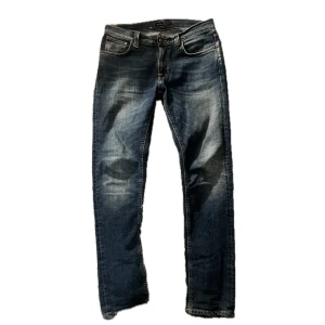 Blå jeans från Nudie Jeans - Snygga blå jeans från Nudie Jeans med klassisk femficksdesign och orangea sömmar på bakfickorna. Jeansen har en rak passform och är tillverkade i bomull med lite stretch för extra komfort. Modellen är Lean Dean och strl 31/32, Skick 9/10
