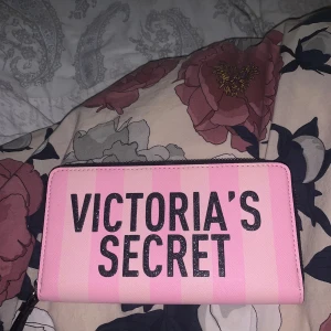 Rosa plånbok från Victoria's Secret - Säljer en rosa plånbok från Victoria's Secret med svart text på framsidan. Plånboken har dragkedja runtom och flera fack för kort och mynt. Perfekt för att hålla ordning på dina värdesaker.