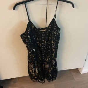 Svart spetsklänning med krokar - Säljer en svart kortklänning med smala axelband och vacker spets över ett beige foder. Klänningen har krokar framtill som detalj och en figurnära passform. Perfekt för dig som vill sticka ut med en elegant och trendig look. Man kan öppna upp knapparna för att visa shortsen eller kjolen beroende på vad man har under. Man måste ha något sånt under annars syns en del av rumpan när man böjer sig