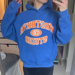 Blå hoodie från WRSTBHVR Studio - Säljer en blå hoodie från WRSTBHVR Studio med orange och vit text framtill. Tröjan har en klassisk huva, känguruficka och ribbade muddar. Så mysig och varm!! Jag har i vanliga fall S och denna sitter lite oversized