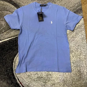 Blå t-shirt från Polo Ralph Lauren - Säljer en klassisk blå t-shirt från Polo Ralph Lauren med rund halsringning och liten vit broderad logga på bröstet. T-shirten är i mjuk bomull och har korta ärmar. Perfekt för en clean och stilren look.