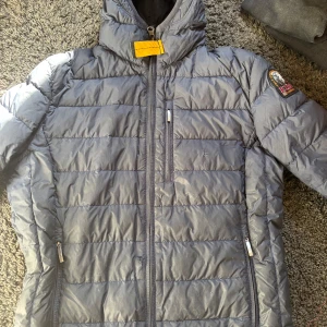 Grå dunjacka från Parajumpers - Säljer en grå dunjacka från Parajumpers med klassisk gul detalj vid kragen och logga på ärmen. Jackan har dragkedja framtill och två sidofickor med dragkedja. Lätt och smidig modell, perfekt för kyliga dagar. Bra skicka bara ett litet hål annars funkar den felfritt. vill ha 1500 kr för den för jag köpte den för 4500 i vintras. pris kan diskuteras inte under 1000.