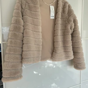Beige fusk pälsjacka från Gina Tricot - Säljer en supermjuk och fluffig beige pälsjacka från Gina Tricot. Jackan har lång ärm och är i kortare modell utan synliga knappar eller dragkedja. Perfekt för att lyfta vilken outfit som helst. Oanvänd och prisettiketen kvar