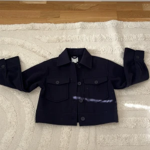 Svart jacka från Gina Tricot - Säljer en svart jacka från Gina Tricot med klassisk krage och stora bröstfickor med knappar. Jackan har långa ärmar och stängs med knappar framtill. Perfekt för dig som gillar en stilren och enkel look. Inte använd. Solen strålar på kappan ingen färg!