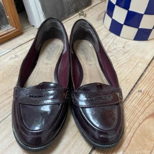 Vinröda loafers  - Säljer ett par vinröda loafers i glansigt lackmaterial. Skorna har klassisk design med rund tå och låg platt sula. Perfekta för dig som gillar stilrena och tidlösa skor med en twist av färg.
