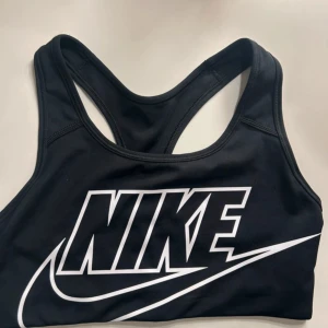 Svart sport-bh från Nike med stor logga - Säljer en svart sport-bh från Nike med stor vit logga framtill. Bh:n är i Dri-Fit-material som andas och har brottarrygg för extra stöd. 
