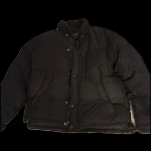 Svart dunjacka från Timberland - Säljer en svart dunjacka från Timberland med hög krage och dragkedja samt knappar framtill. Jackan har en klassisk pufferstil och är perfekt för kyliga dagar. Enkel och stilren design som passar till det mesta. Skönt å varmt material, den är även vintage köpt under 80-2000s talet 