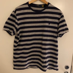 Randig t-shirt från Polo Ralph Lauren - Säljer en klassisk randig t-shirt från Polo Ralph Lauren i mörkblått och grått. T-shirten har korta ärmar, rund hals och den ikoniska broderade loggan på bröstet. Perfekt för en stilren och avslappnad look.