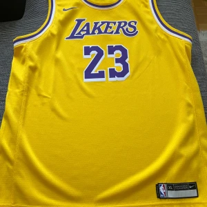 Gul Lakers baskettröja med nummer 23 - Säljer en gul Los Angeles Lakers baskettröja från Nike med nummer 23 och namnet James på ryggen. Tröjan har lila och vita detaljer, är ärmlös och tillverkad i Dri-FIT-material för bra ventilation. Perfekt för basketfans!