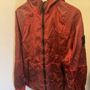 Röd vindjacka från Stone Island - Säljer en röd vindjacka från Stone Island med huva och svarta dragkedjor. Jackan har märkets klassiska patch på ärmen och är tillverkad i ett lätt syntetmaterial. Perfekt för blåsiga dagar och enkel att packa ner i väskan.