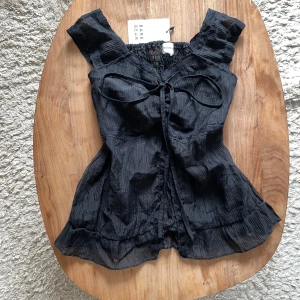 NY y2k fairy blus med volang och knyt - Supersöt svart blus från Bare Dare med volangdetaljer och knyt framtill🎀 Blusen har breda axelband, smockad rygg och lätt rynkad nederkant. Är figursydd.