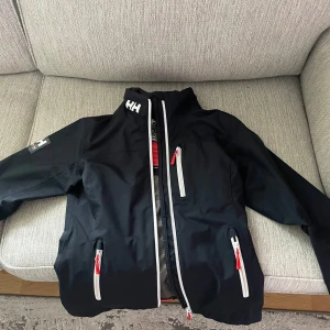 Svart vindjacka från Helly Hansen - Svart vindjacka från Helly Hansen med vita och röda detaljer på dragkedjorna.  Storlek S bra skick
